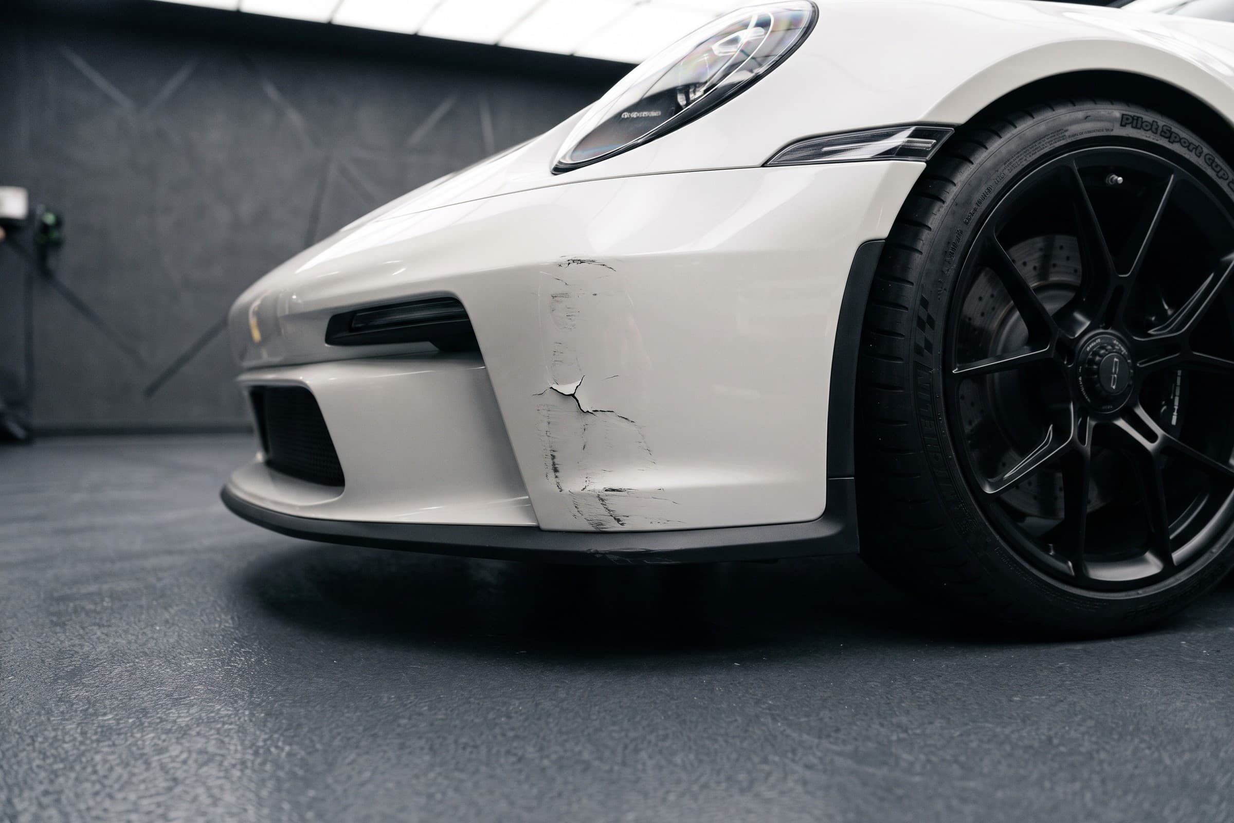 Close-up of an existing crack and marks on a white bumper, illuminated inside a car detailing studio.